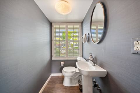 Bright and modern half bathroom featuring a sleek pedestal sink, a round mirror, and natural light from the window with shutters. Perfect for a quick refresh in a stylish and functional space.