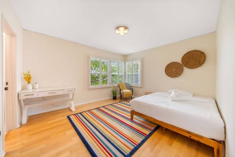 Bright and airy bedroom with cozy beds, vibrant rug, and charming decor. Features a writing desk and large windows for natural light, creating a relaxing and inviting space for rest or work.